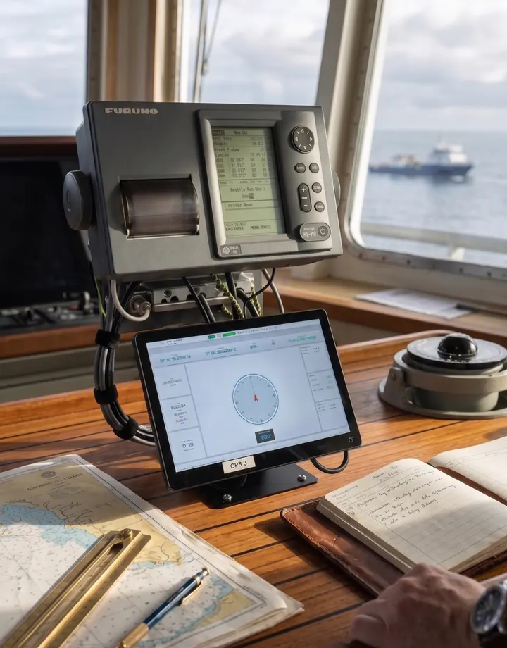 DIDI GNSS display on a wheelhouse chart desk beside a Furuno GNSS receiver, a paper chart and the deck log